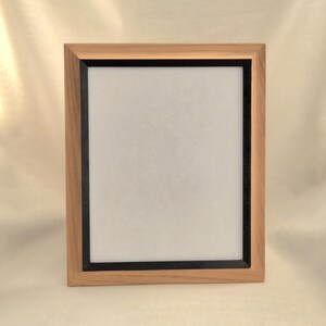 May include: A rectangular wooden picture frame with a light brown finish. The frame has a black inner border and a white interior, suitable for displaying a photograph or artwork. The frame is designed to be displayed on a wall or tabletop.