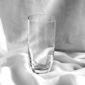 May include: A clear, tall drinking glass with a slightly flared rim. The glass has a simple, elegant design and sits on a white, textured surface. The glass is empty and appears to be made of crystal or high-quality glass.