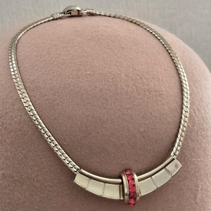 May include: Silver-toned necklace with a textured chain and a curved centerpiece. The centerpiece features a band of pink gemstones and rectangular silver accents. The necklace is displayed on a pink surface.