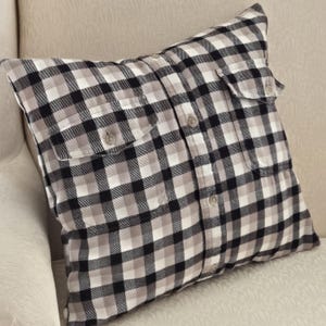 May include: A square pillow made from a black, white, and gray plaid flannel shirt. The pillow has two buttoned pockets and buttons down the front. The pillow is resting on a cream-colored armchair.