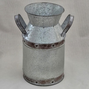 May include: A galvanized metal milk can-style vase with two handles. The vase has a flared top and a cylindrical body with a rusted brown band around the top and bottom. The vase is approximately 10 inches tall.