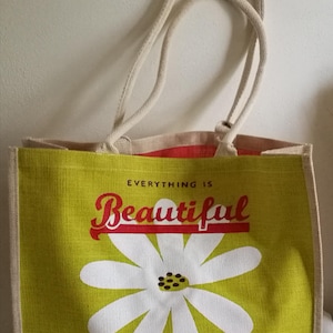 May include: A green tote bag with cream-colored handles and a white daisy graphic. The bag features the text "Everything is Beautiful in its Time" in red and white. The scripture reference Isaiah 61:3 is also included.