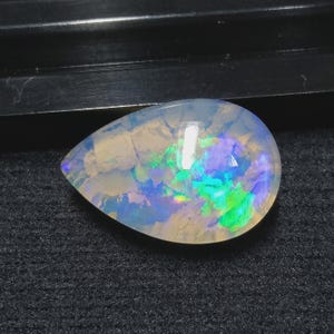 Può includere: Una gemma di opale a forma di goccia con una base bianca lattea traslucida. La pietra presenta lampi interni di colore iridescente, tra cui blu, verdi e viola. L'opale è posto su uno sfondo scuro.