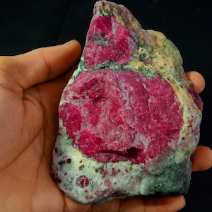 Puede incluir: Una gran gema de rubí rosa en bruto con inclusiones verdes y blancas. El rubí está en una mano, mostrando su tamaño y forma.