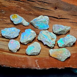 Peut inclure: Une collection de dix pierres d'opale brutes avec des couleurs irisées bleues, vertes et orange. Les pierres sont de tailles et de formes variées et ont une matrice blanche, grise et brune.