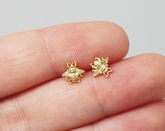 May include: A pair of small, gold-colored bee-shaped stud earrings. The earrings are detailed with textured wings and bodies, and are displayed on a light background. The earrings are small and delicate.