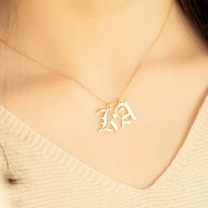 Puede incluir: Un collar dorado con dos letras colgantes en una fuente inglesa antigua. El collar se lleva alrededor del cuello, y los colgantes son visibles contra una parte superior de color claro. Las letras parecen ser "KA".