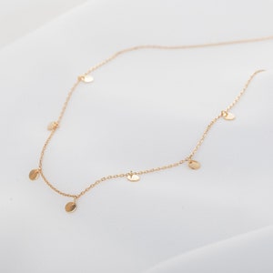 Peut inclure: Délicat collier en or avec de petits disques ronds espacés le long de la chaîne. Le collier est présenté sur un tissu blanc texturé, soulignant son élégance simple et son design minimaliste.