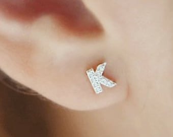 May include: A close-up of an ear with a small, silver-colored stud earring in the shape of the letter "K". The earring is encrusted with small, clear gemstones, adding sparkle. The skin around the ear is a light, neutral tone.
