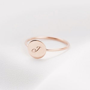 May include: A rose gold ring with a circular face. The face of the ring has the letter "J" engraved in a delicate script. The ring is set against a white, silky background.