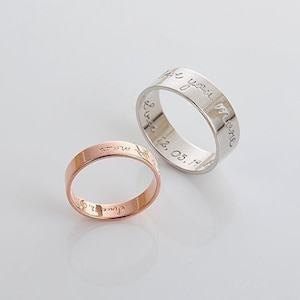 Puede incluir: Dos anillos, uno de oro rosa y otro de plata, con texto grabado. El anillo de oro rosa tiene una banda plana y el anillo de plata tiene una banda más ancha. Los anillos están sobre una superficie blanca.