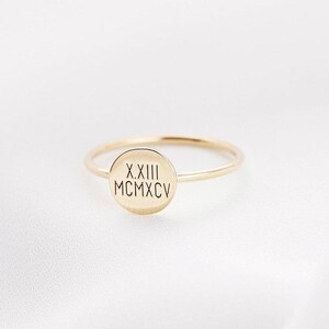 May include: A delicate gold ring with a thin band and a circular face. The face of the ring features engraved Roman numerals. The ring is set against a white background.