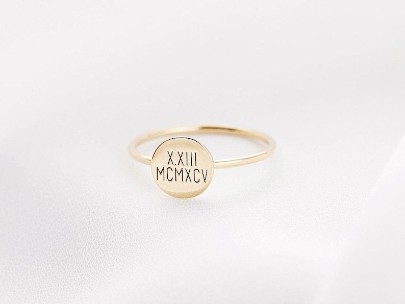 May include: A delicate gold ring with a thin band and a circular face. The face of the ring features engraved Roman numerals. The ring is set against a white background.