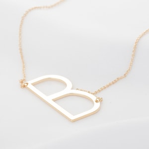 Puede incluir: Collar dorado con un colgante con la letra "B". El colgante es un diseño grande y abierto, suspendido de una delicada cadena. El collar se muestra sobre un fondo blanco, resaltando la simple elegancia de la joya.