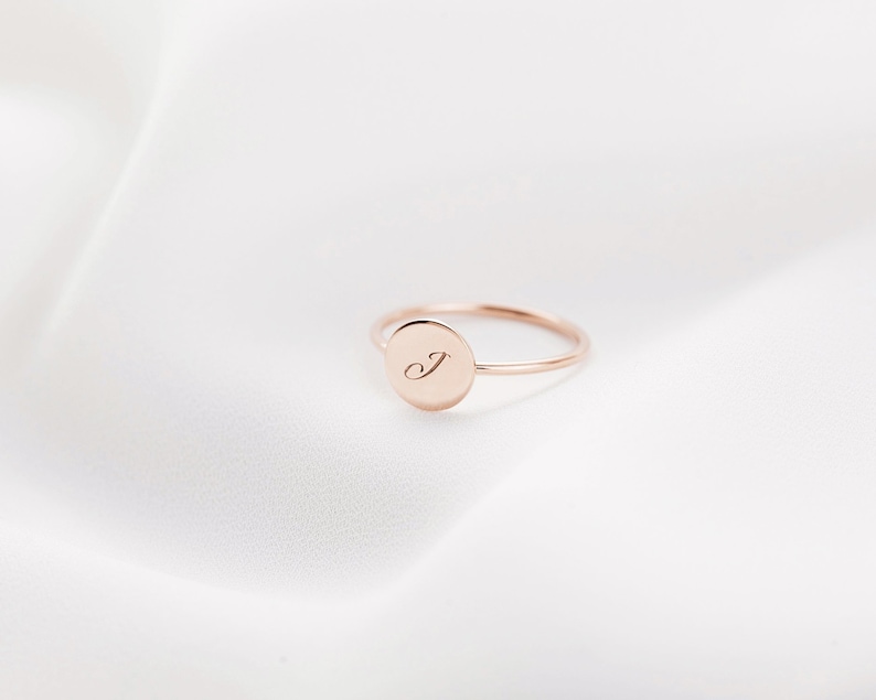 May include: A rose gold ring with a circular face. The face of the ring has the letter "J" engraved in a delicate script. The ring is set against a white, silky background.
