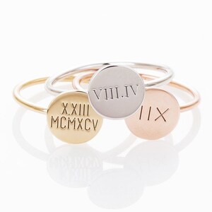 May include: A collection of personalized rings in gold, silver, and rose gold. Each ring features a circular disc engraved with Roman numerals. The rings are displayed on a white surface, showcasing their polished finish and delicate design.