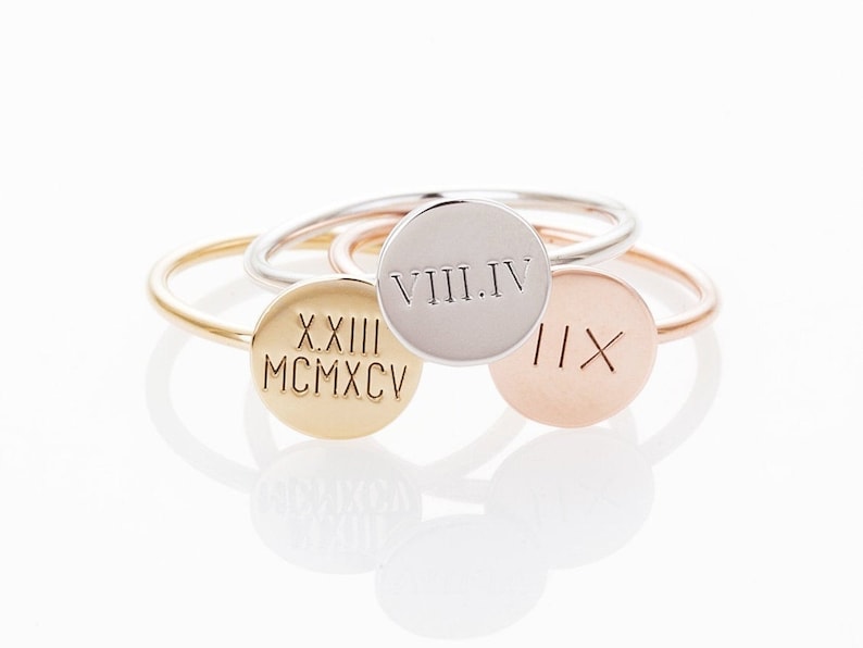 May include: A collection of personalized rings in gold, silver, and rose gold. Each ring features a circular disc engraved with Roman numerals. The rings are displayed on a white surface, showcasing their polished finish and delicate design.