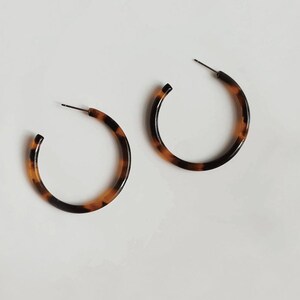 May include: A pair of hoop earrings with a tortoiseshell pattern. The earrings are a warm brown color with dark brown accents. The hoops are round and have a post for insertion into the ear.