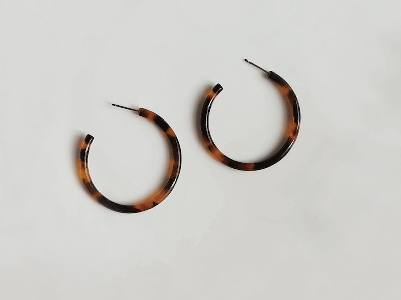 May include: A pair of hoop earrings with a tortoiseshell pattern. The earrings are a warm brown color with dark brown accents. The hoops are round and have a post for insertion into the ear.