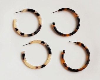 May include: Four pairs of hoop earrings in a tortoiseshell pattern. Two pairs have a light beige and black pattern, and the other two pairs have a brown and black pattern. The earrings have a post closure.