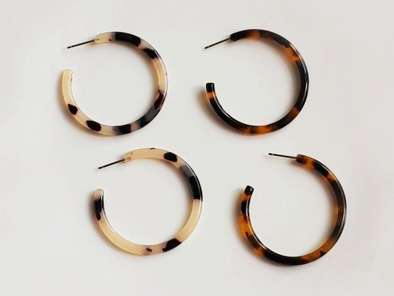 May include: Four pairs of hoop earrings in a tortoiseshell pattern. Two pairs have a light beige and black pattern, and the other two pairs have a brown and black pattern. The earrings have a post closure.