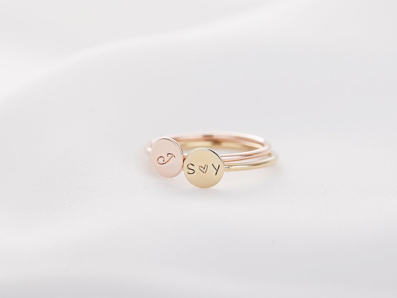 May include: Three delicate rings in rose gold, yellow gold, and gold. Each ring features a small, round charm with engraved initials and a heart symbol. The rings are displayed on a white, textured surface.