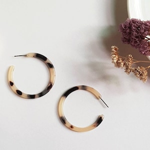 May include: Two hoop earrings with a tortoiseshell pattern. The earrings are a mix of cream, brown, and black. The earrings are on a white surface with a white mug and dried flowers in the background.