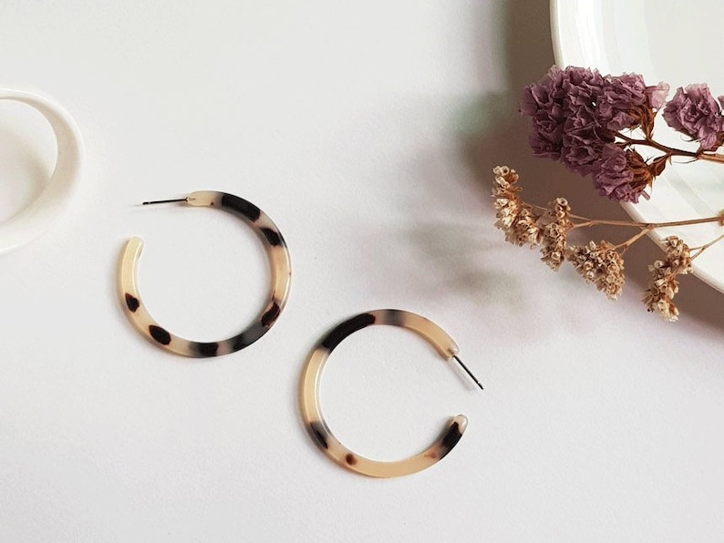 May include: Two hoop earrings with a tortoiseshell pattern. The earrings are a mix of cream, brown, and black. The earrings are on a white surface with a white mug and dried flowers in the background.