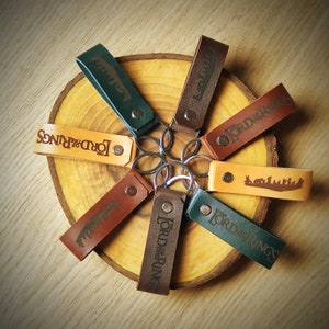 May include: A set of seven leather keychains featuring the Lord of the Rings logo and various designs. The keychains are arranged in a circle on a wooden surface.