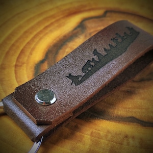 May include: A brown leather keychain with a silhouette of a group of people walking on a path. The keychain has a silver metal ring.