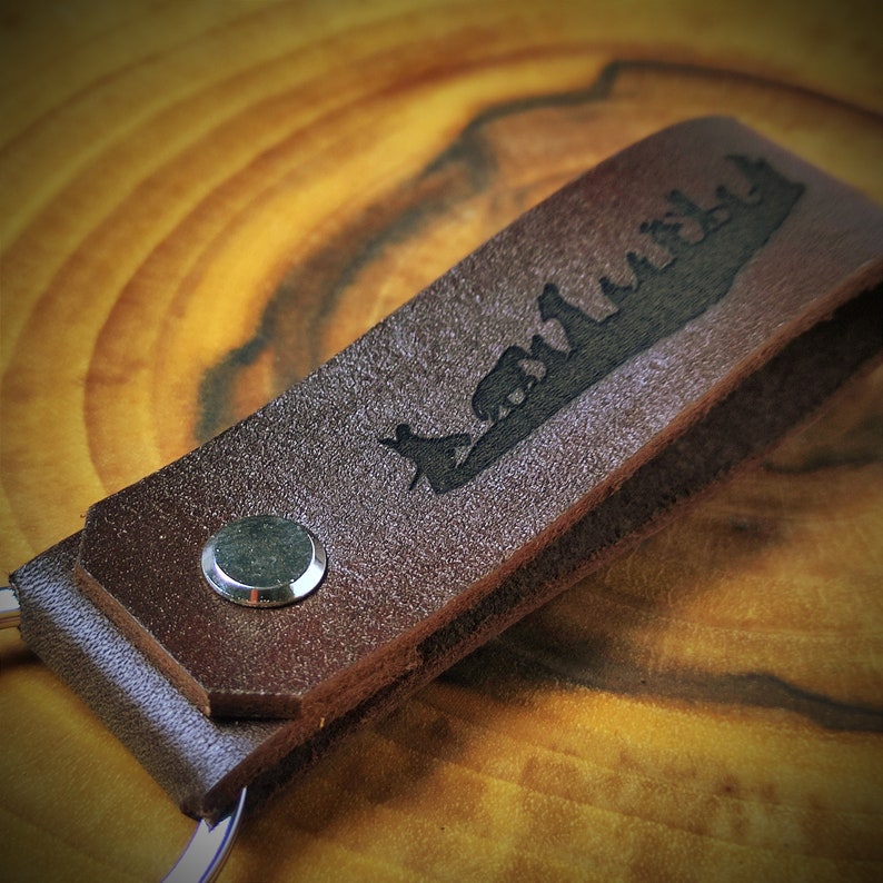 May include: A brown leather keychain with a silhouette of a group of people walking on a path. The keychain has a silver metal ring.