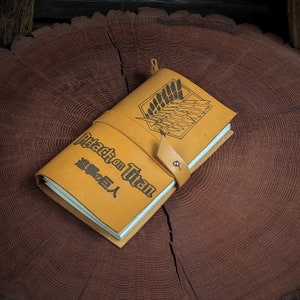May include: A brown leather journal with the words "Attack on Titan" and Japanese characters on the cover. The journal has a strap closure and a leather wing design.