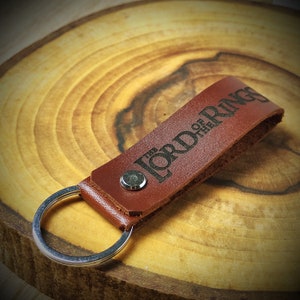 Puede incluir: Un llavero de cuero marrón con un anillo de llave plateado. El llavero tiene las palabras "The Lord of the Rings" grabadas en relieve.