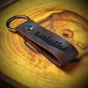 May include: A brown leather keychain with a silver ring and a black silhouette of a forest on it.