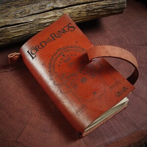 May include: A brown leather-bound journal with a decorative design of the entrance to the Mines of Moria from "The Lord of the Rings" and the text "The Lord of the Rings" on the cover. The journal has a leather strap closure and the initials "D.B." are embossed on the back cover.