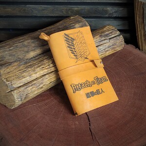 May include: A brown leather notebook with the text "Attack on Titan" and the Japanese text "進撃の巨人" printed on the cover. The cover also features a black and white graphic of the Wings of Freedom symbol.