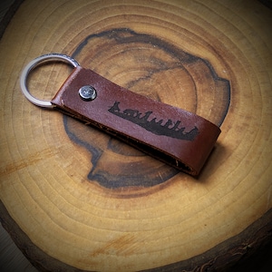 Puede incluir: Un llavero de cuero marrón con un anillo de llave plateado. El llavero tiene una silueta de un grupo de personas grabada en el cuero.