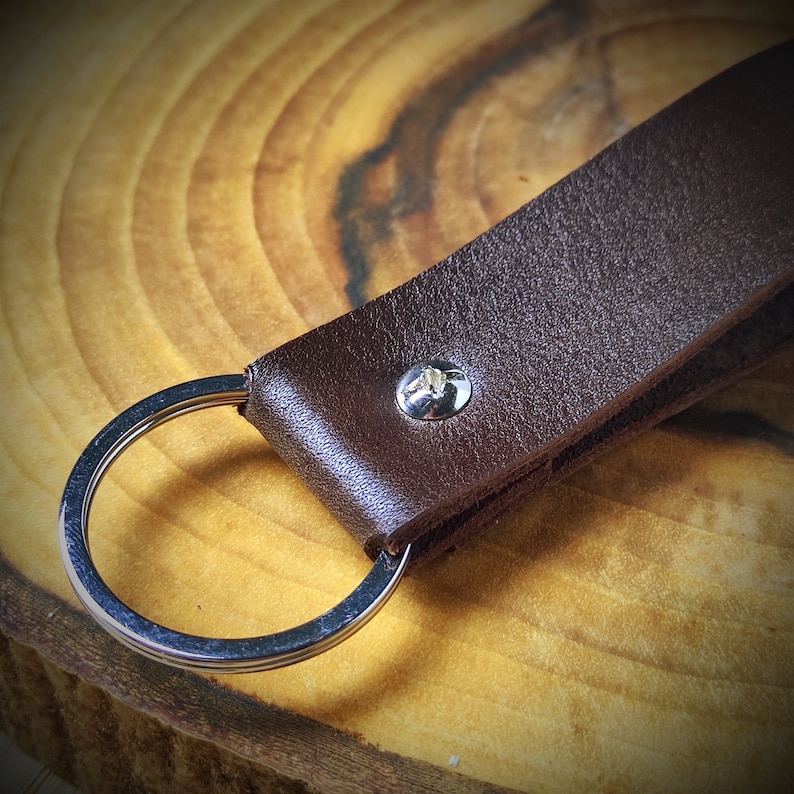 May include: A brown leather keychain with a silver metal ring. The keychain has a silver metal screw in the center.