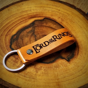 Puede incluir: Un llavero de cuero marrón con un anillo de llave plateado. El llavero tiene las palabras "The Lord of the Rings" grabadas en relieve.