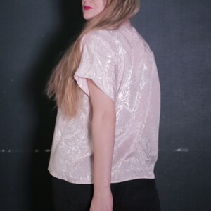 Pode incluir: Uma blusa rosa de manga curta com um padrão sutil em relevo. A blusa tem um caimento solto e uma gola ligeiramente franzida.