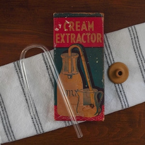 May include: An antique cream extractor set with a glass tube, a wooden stopper, and a vintage box. The box has a red top with the words "Cream Extractor" and an illustration of a milk bottle and pitcher.