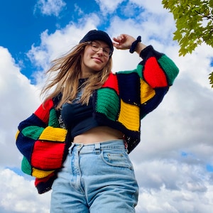 Könnte beinhalten: Ein farbenfroher Patchwork-Häkelcardigan in Rot, Grün, Gelb und Marineblau. Der Cardigan wird mit einem schwarzen Oberteil und hellblauen Jeans getragen. Eine schwarze Mütze und eine Brille vervollständigen den Look. Der Hintergrund zeigt einen blauen Himmel mit Wolken.