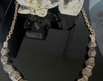 May include: A gold chain necklace with a large, chunky pyrite bead pendant. The beads are faceted and have a dark, metallic sheen. The necklace is displayed on a black surface with a clear crystal lotus flower and a clear crystal point.