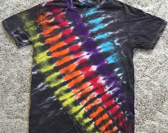 May include: A dark gray t-shirt with a vibrant tie-dye pattern. The design features diagonal stripes of yellow, orange, red, purple, and blue. The shirt has short sleeves and a crew neck, perfect for casual wear.