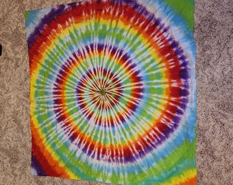 May include: A square, tie-dye textile with a spiral pattern. The design features concentric rings of color, including red, orange, yellow, green, blue, and purple. The fabric is likely cotton or a similar material, with a soft, slightly wrinkled texture.