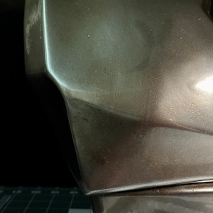 May include: Close-up of a metallic grey helmet with a textured surface. The helmet has a sharp, angular design.