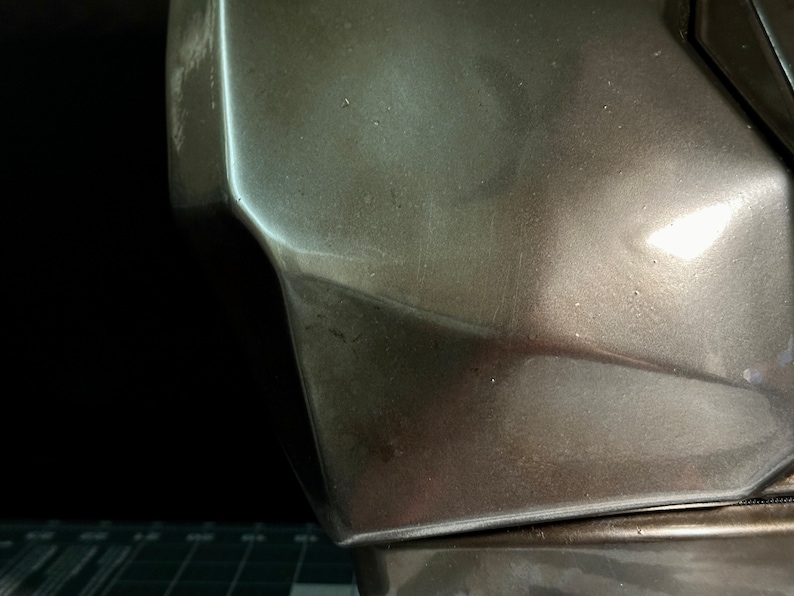 May include: Close-up of a metallic grey helmet with a textured surface. The helmet has a sharp, angular design.