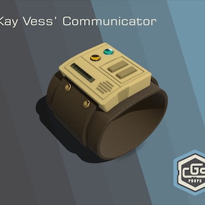 Puede incluir: Un comunicador de muñeca beige y marrón con una pantalla digital, dos botones y una luz amarilla y verde. El texto "Kay Vess' Communicator" está por encima del dispositivo. El dispositivo está en una correa de cuero marrón.