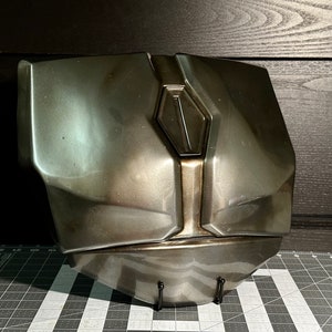 May include: A metallic grey and silver chest piece with a raised, diamond-shaped design in the centre. The chest piece is on a black stand.