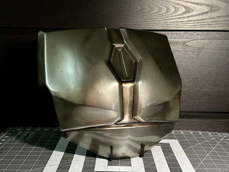 May include: A metallic grey and silver chest piece with a raised, diamond-shaped design in the centre. The chest piece is on a black stand.